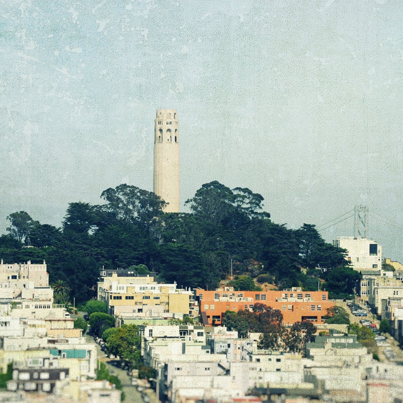 May include: A view of the Coit Tower in San Francisco, California, with a cityscape in the foreground. The tower is a white, cylindrical structure with a pointed top. The cityscape is a mix of residential buildings and trees.