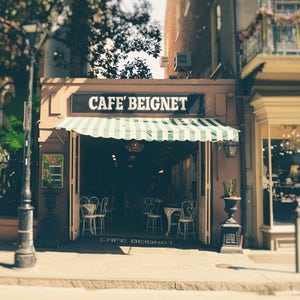 Op de afbeelding: Een caf&eacute; met een groen-wit gestreept luifel en de tekst "Cafe Beignet" op het bord. Het caf&eacute; heeft een vintage uitstraling en bevindt zich in een straat met andere gebouwen op de achtergrond.