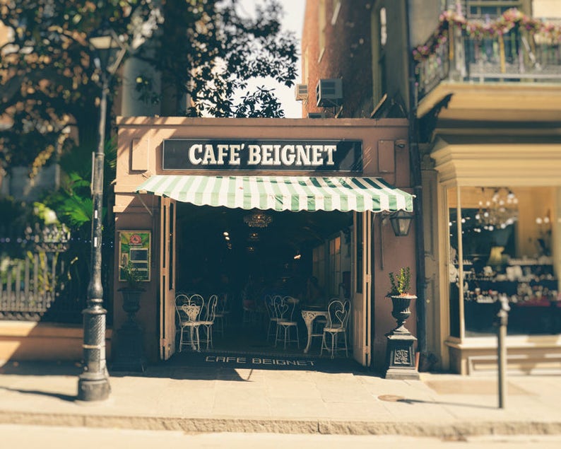 Op de afbeelding: Een caf&eacute; met een groen-wit gestreept luifel en de tekst "Cafe Beignet" op het bord. Het caf&eacute; heeft een vintage uitstraling en bevindt zich in een straat met andere gebouwen op de achtergrond.