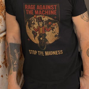 May include: Black t-shirt with a retro graphic. The design features the text "RAGE AGAINST THE MACHINE" above an illustration of a monkey on a unicycle juggling balls, with the words "STOP THE MADNESS" below.
