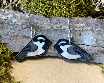 May include: A pair of silver earrings featuring a black and white painted bird design. The birds are facing to the right and have a white belly and black wings and head.