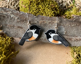 May include: A pair of dangle earrings featuring hand-painted black-capped chickadee birds on a white background. The earrings are made of wood and have silver hooks.