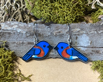 May include: A pair of bluebird earrings made of painted wood with silver hooks. The birds are blue with a red breast and white accents.