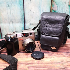 May include: A silver Olympus Camedia C-2040 Zoom digital camera with a black lens cap and a black leather camera bag. The camera has a black strap attached to it.