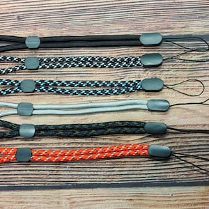 May include: An overhead shot shows six wrist straps in different colours and patterns. The straps are black, black and white, blue and black, grey, black and green, and red and white. Each strap has a loop and a small black cord.