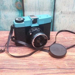 May include: A vintage Diana F+ camera. It has a black body and a light blue top. The camera features a black lens, a black strap, and a lens cap. The lens cap is printed with the words "DIANA F+".