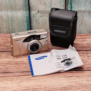 May include: A silver Samsung Rocas 110 Impax film camera with a black carrying case and instruction manual. The camera has a lens cover and a silver Samsung logo.