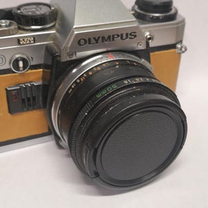 May include: A vintage Olympus camera with a 50mm lens and lens cap. The camera body is silver and tan, with black dials and a black lens. The lens has white markings. The word "OLYMPUS" is printed on the top of the camera.