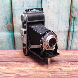 Peut inclure: Un appareil photo pliable noir et argenté vintage avec une poignée en cuir et un soufflet. L'objectif est argenté avec des marquages noirs. L'appareil photo est ouvert, révélant l'objectif et les composants internes. L'appareil photo est sur une surface en bois.