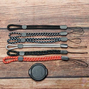 May include: A collection of five camera wrist straps in various colors and patterns, including black, white and black checkered, blue and black checkered, gray, black and green camouflage, and red and white. A black lens cap is also present.