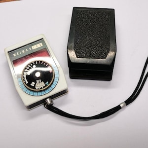 May include: A vintage Weimar Lux light meter with a black and white dial, a white body, and a black neck strap. A black case with a textured top is next to the meter. The meter has a circular dial with numbers and markings.