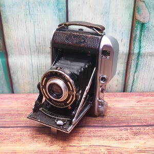 May include: A vintage black and silver folding camera with a brown leather handle. The camera has a Prontor lens and is open, revealing the bellows. The camera is on a wooden surface.