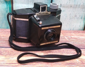 Puede incluir: Una cámara Flashmaster 35 mm Coronet negra con una correa negra. La cámara tiene una lente plateada y una placa de identificación plateada que dice "Flashmaster 35 mm Coronet Made in England".