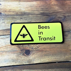 Puede incluir: Un letrero amarillo con texto negro y un símbolo de advertencia triangular negro. El letrero dice "Bees in Transit" y el triángulo contiene una silueta de una abeja.