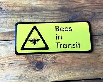 May include: A yellow sign with black text and a black triangle warning symbol. The sign reads "Bees in Transit" and the triangle contains a silhouette of a bee.