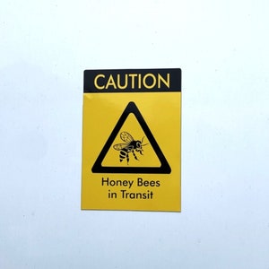 May include: Yellow and black caution sign with a black triangle and a bee inside. The sign reads "CAUTION Honey Bees in Transit".