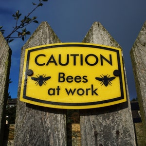 May include: A yellow sign with black text that reads "CAUTION Bees at work" with two black bee silhouettes on either side of the text. The sign is attached to a wooden fence.