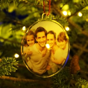 Puede incluir: Un adorno navideño redondo transparente con una foto de una familia de cuatro. La foto muestra un hombre, una mujer y dos niños. El adorno está colgado de un árbol de Navidad.