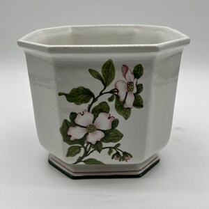 May include: A white ceramic planter with an octagonal shape and a painted floral design featuring pink dogwood blossoms and green leaves. The base of the planter is accented with a thin red and green line, and the interior is plain white.