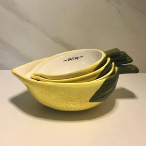 May include: A set of four ceramic measuring cups shaped like lemons. The cups are yellow with green leaf accents and feature the text "- 1/4 Cup -". The cups are nested together, with the smallest cup visible.