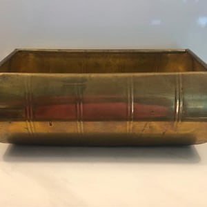 May include: A rectangular brass planter with a polished metallic finish and rounded bottom edges. The exterior surface features vertical embossed lines spaced evenly along the length of the container, creating a structured and uniform appearance.