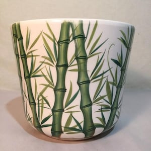 May include: White ceramic planter with a green bamboo design. The planter has a wide mouth and a rounded bottom.