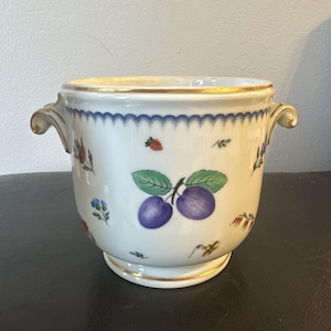 Puede incluir: Un cubo de hielo de porcelana blanca con ribetes dorados y un patrón floral con ciruelas, hojas y flores. El cubo tiene dos asas.