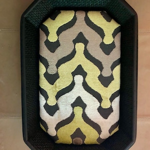 May include: A rectangular pet bed with a black woven frame. The bed's cushion features a repeating pattern of yellow, gray, and white shapes on a black background. The bed is designed for pets.