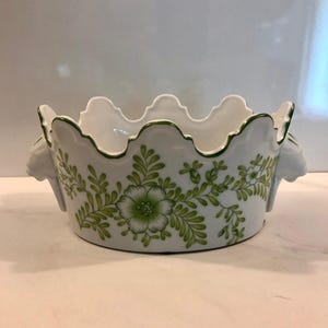 May include: White ceramic bowl with a scalloped rim and green floral design. The bowl features a repeating pattern of green flowers and leaves. Two small handles are on either side of the bowl. The rim is outlined in green.