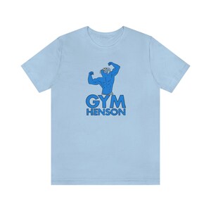 Puede incluir: Camiseta azul claro con una ilustración de dibujos animados de una figura azul musculosa con cabeza de pájaro flexionando los bíceps. El texto "GYM HENSON" está impreso debajo de la figura.