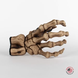 Może przedstawiać: Ceramiczna ręka szkieletowa z brązowo-białym wykończeniem. Ręka jest ułożona z wyciągniętymi i lekko zgiętymi palcami. Ręka jest przedmiotem dekoracyjnym.