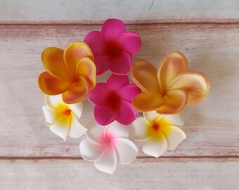 Puede incluir: Seis flores de plumeria artificiales en varios colores, incluyendo amarillo, rosa y blanco, dispuestas sobre un fondo de madera clara.