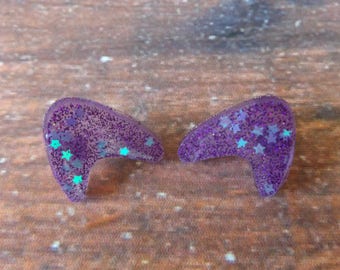 May include: A pair of purple resin earrings shaped like boomerangs with iridescent glitter and small blue star accents.