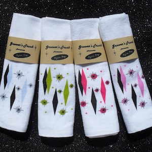 May include: Four white kitchen towels with a retro design featuring black diamonds, white stars, and colored accents. The towels are labeled "Groover's Trunk" with the text "Atomic" and "Pigeon Bay".
