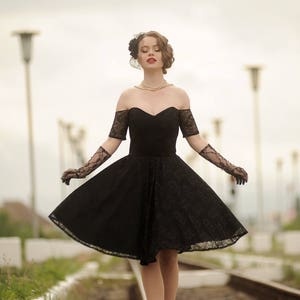 May include: A woman in a black lace dress and gloves walks on a railroad track. She is wearing a pearl necklace and a black veil.