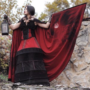 May include: A woman in a gothic-style costume stands on a stone wall, holding a lantern. The outfit features a red and black dress with a matching cape. The cape has a dark design and is spread out, creating a dramatic effect.