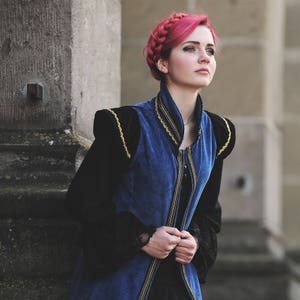 May include: A woman with pink braided hair wearing a blue and black medieval-style vest with gold trim. The vest has a high collar and is buttoned up the front. The sleeves are black and the background is a stone wall.