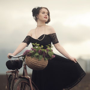 May include: A woman in a black lace dress and a pearl necklace rides a vintage bicycle with a wicker basket filled with purple flowers.