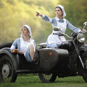 Puede incluir: Dos mujeres vestidas de enfermeras van en una motocicleta negra con sidecar. La mujer en el sidecar mira hacia un lado, mientras que la mujer que conduce la motocicleta apunta hacia la derecha. La motocicleta est&aacute; en un campo de hierba con un fondo borroso de &aacute;rboles.