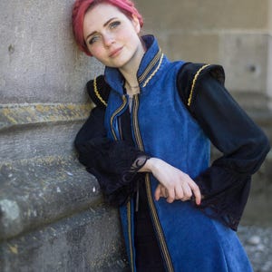 May include: A blue vest with gold trim and black sleeves. The vest has a high collar and a zipper down the front. The sleeves are long and black with lace detailing. The person is leaning against a stone wall.
