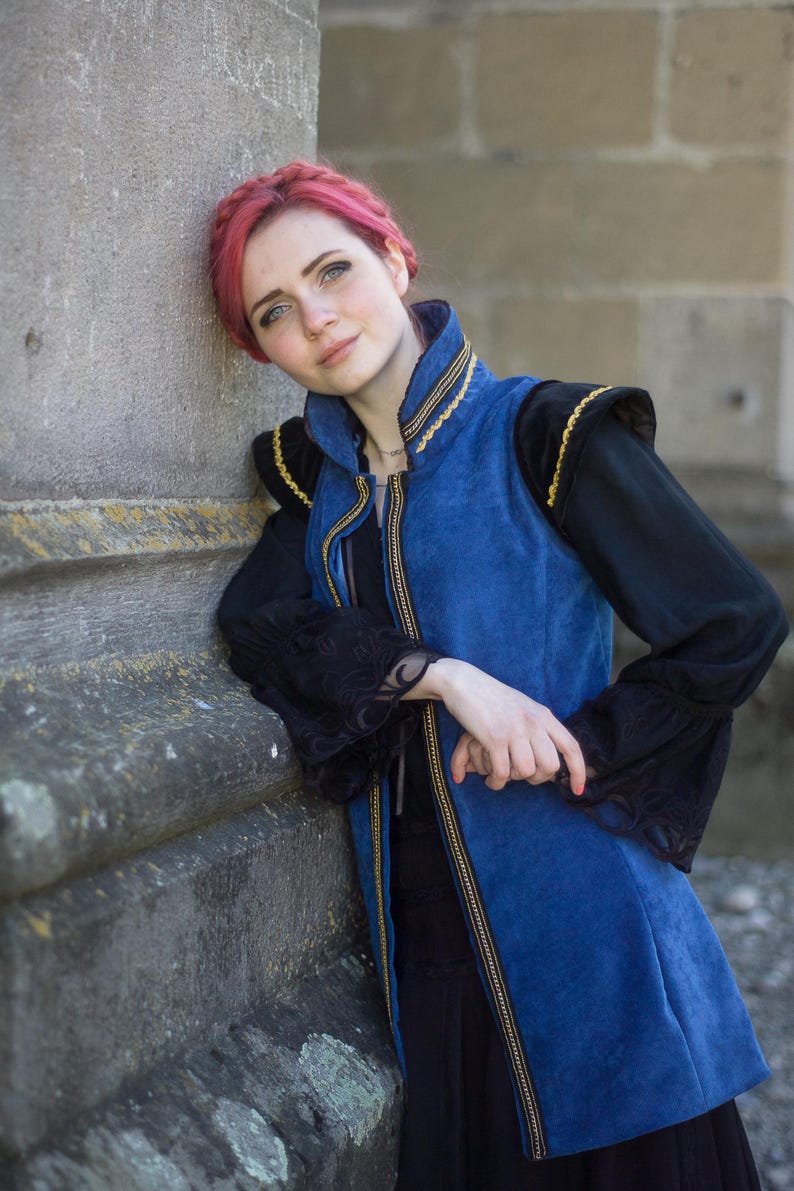 May include: A blue vest with gold trim and black sleeves. The vest has a high collar and a zipper down the front. The sleeves are long and black with lace detailing. The person is leaning against a stone wall.