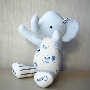 May include: A light blue plush elephant toy with a white belly featuring blue dog illustrations. The elephant has striped gray and white legs and ears. The name "Carol" is embroidered on one foot, and "Teodor" on the other.