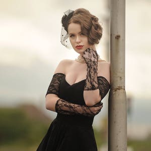 May include: A woman in a black off-the-shoulder dress with lace detailing and a black lace glove. She is wearing a pearl necklace and a black veil headband.