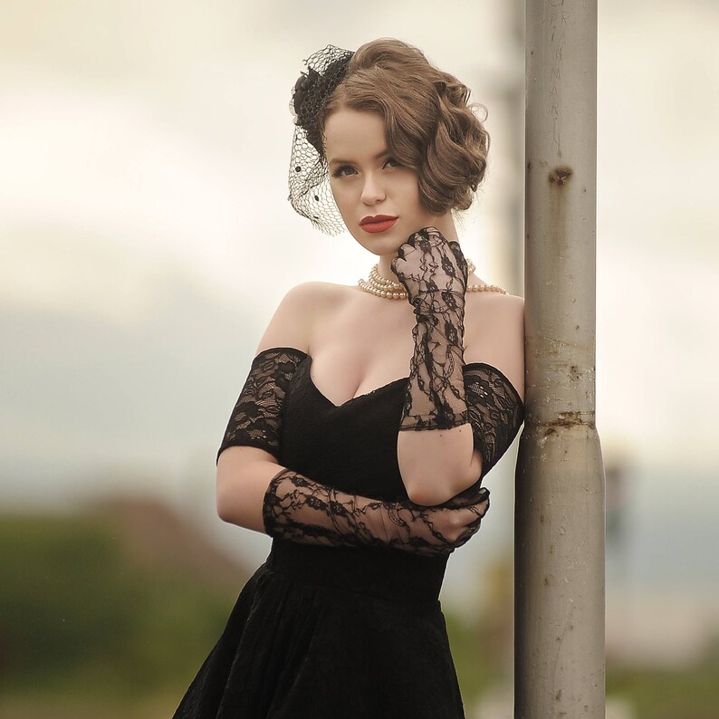 May include: A woman in a black off-the-shoulder dress with lace detailing and a black lace glove. She is wearing a pearl necklace and a black veil headband.