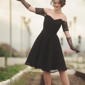 May include: A woman in a black lace dress and black gloves walks on a set of train tracks. She is wearing a pearl necklace and a black hat with a veil.