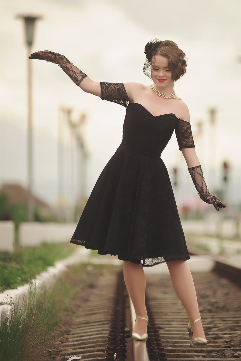 May include: A woman in a black lace dress and black gloves walks on a set of train tracks. She is wearing a pearl necklace and a black hat with a veil.