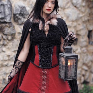 May include: A woman in a black cape and a red and black dress holds a lantern. The dress features a red skirt with black lace trim and a black bodice with ornate detailing. The cape has a fur collar.