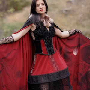 May include: A gothic-style dress with a red skirt and a black bodice. The dress features intricate lace and beadwork details. A matching red cape with a black design is draped over the shoulders. The outfit is completed with lace gloves and a fur collar.