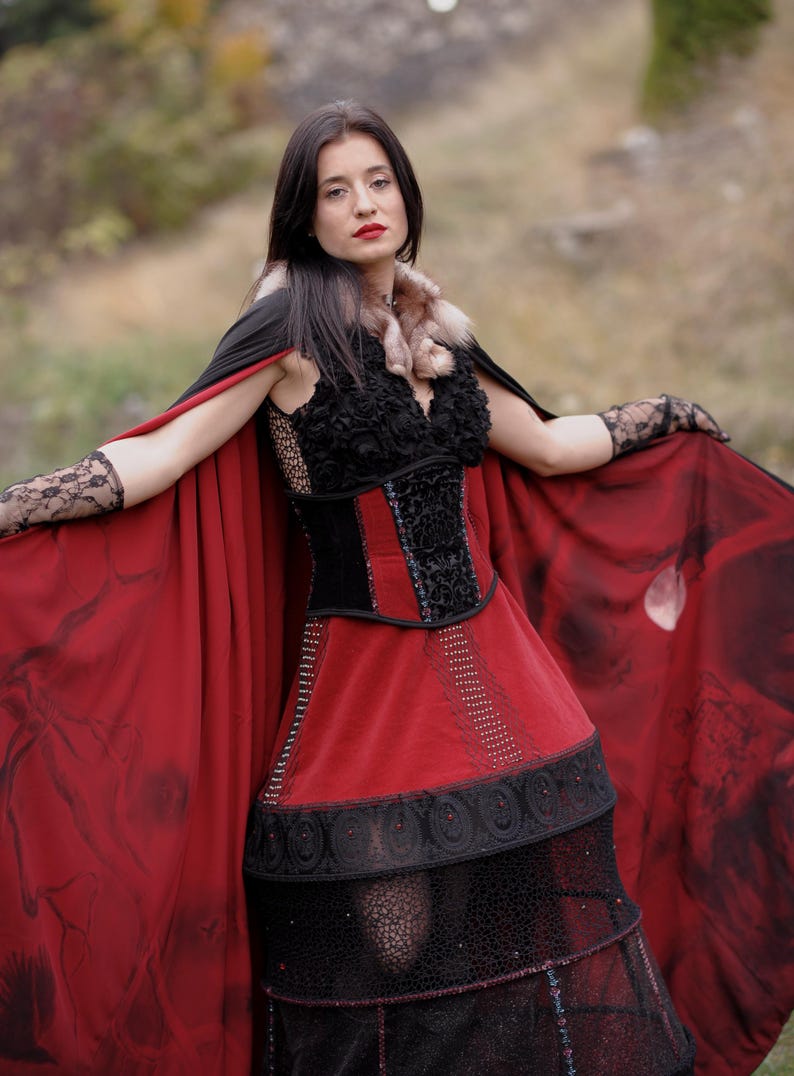 May include: A gothic-style dress with a red skirt and a black bodice. The dress features intricate lace and beadwork details. A matching red cape with a black design is draped over the shoulders. The outfit is completed with lace gloves and a fur collar.