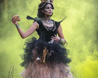 May include: A person in a dark fantasy costume with a skull headdress and holding a skull. The outfit includes a black bodice, layered tulle skirt, and decorative bones. The background is a hazy yellow.